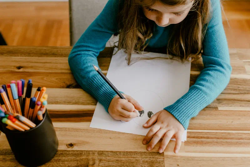 Menina desenhando em uma mesa de madeira, ao lado de um pote de lápis de cor
