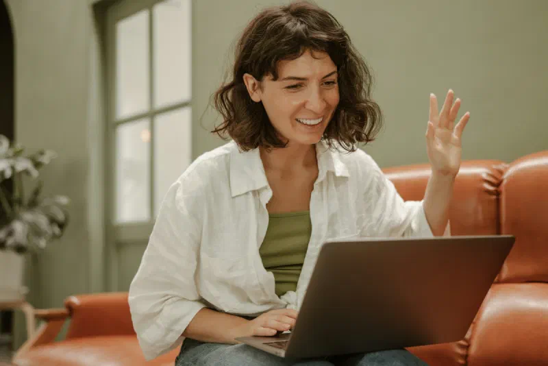 Pessoa sorrindo e acenando para uma chamada de vídeo no notebook, sentada no sofá