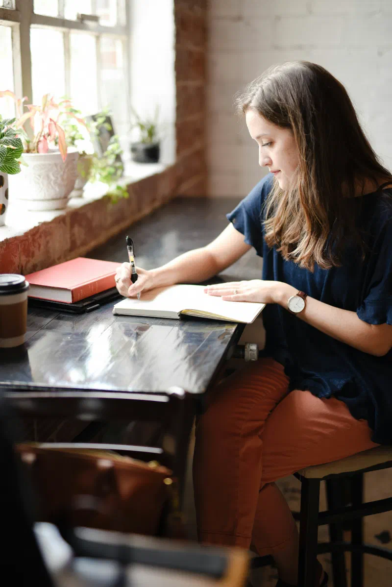 Mulher escrevendo em um caderno, sentada a uma mesa perto de uma janela com plantas
